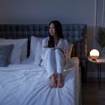 A woman in pajamas sits on a bed in a dimly lit bedroom, creating a serene nighttime atmosphere.