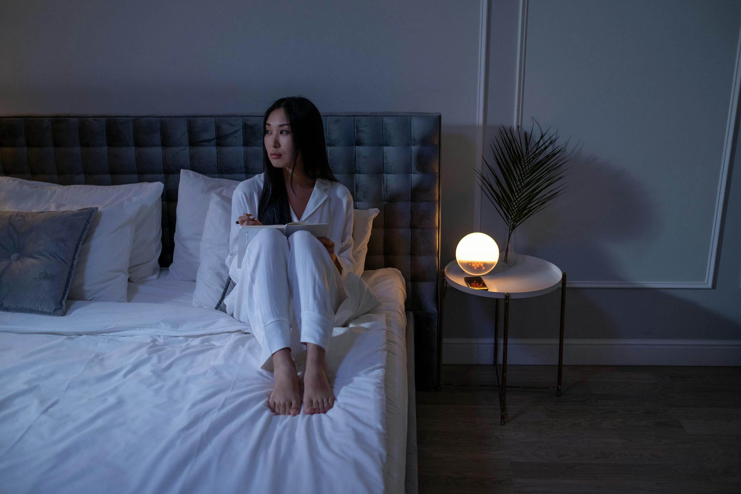 A midlife woman in pajamas sits on a bed in a dimly lit bedroom, creating a serene nighttime atmosphere.