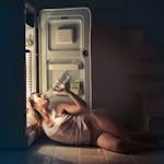 A perimenopause woman in sleepwear drinking water beside an open fridge late at night, conveying thirst and refreshment.