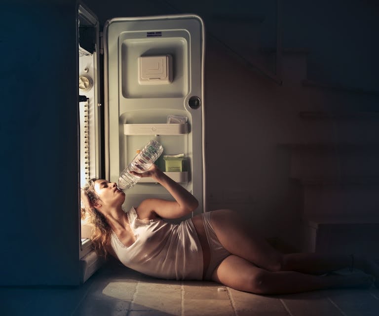 A perimenopause woman in sleepwear drinking water beside an open fridge late at night, conveying thirst and refreshment.