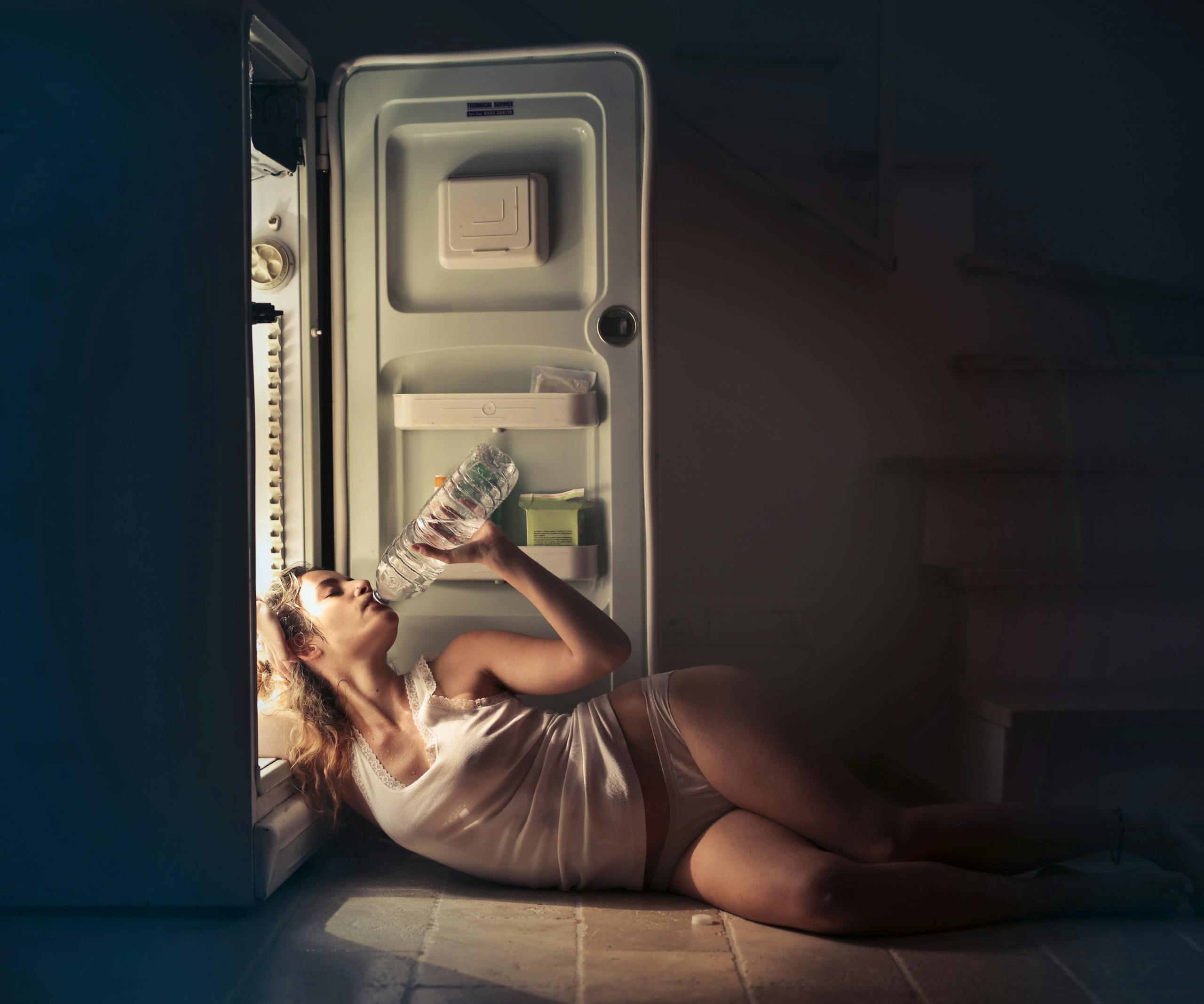 A perimenopause woman in sleepwear drinking water beside an open fridge late at night, conveying thirst and refreshment.