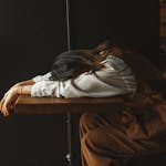 A perimenopausal woman in casual attire rests her head on a wooden table in a dimly lit room, expressing fatigue.