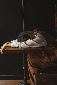 A perimenopausal woman in casual attire rests her head on a wooden table in a dimly lit room, expressing fatigue.