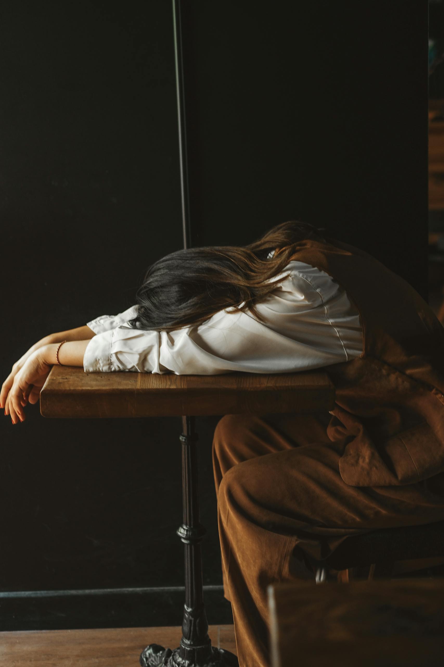 A perimenopausal woman in casual attire rests her head on a wooden table in a dimly lit room, expressing fatigue.