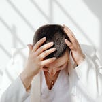 Perimenopausal woman in white outfit feeling tired and stressed after not enough sleep, sitting with hands on head in bright sunlight indoors.