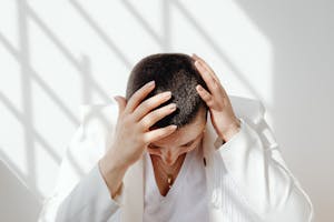 Perimenopausal woman in white outfit feeling tired and stressed after not enough sleep, sitting with hands on head in bright sunlight indoors.