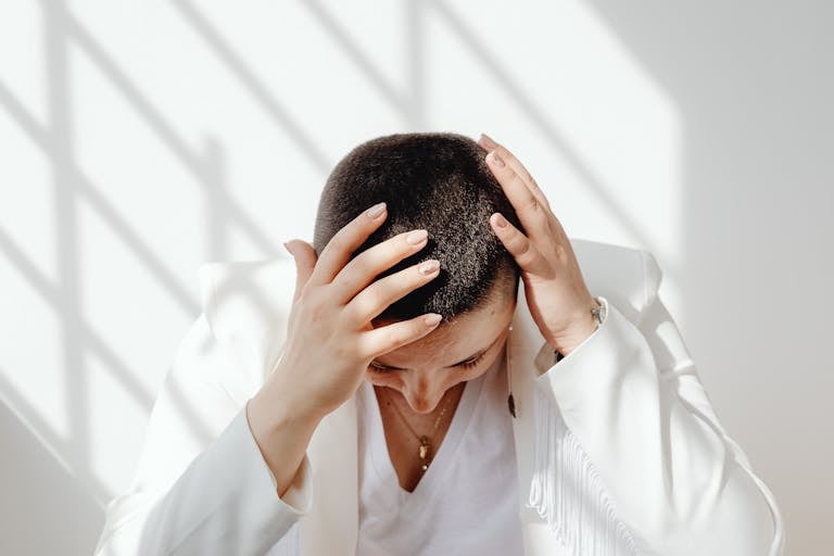 Perimenopausal woman in white outfit feeling tired and stressed after not enough sleep, sitting with hands on head in bright sunlight indoors.