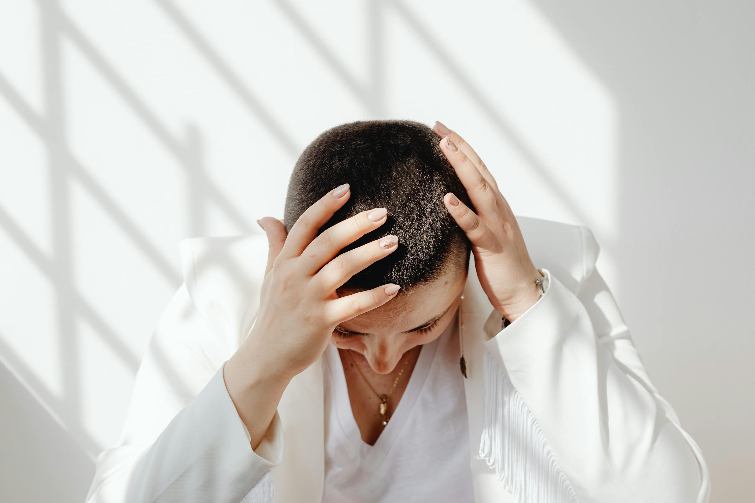 Perimenopausal woman in white outfit feeling tired and stressed after not enough sleep, sitting with hands on head in bright sunlight indoors.
