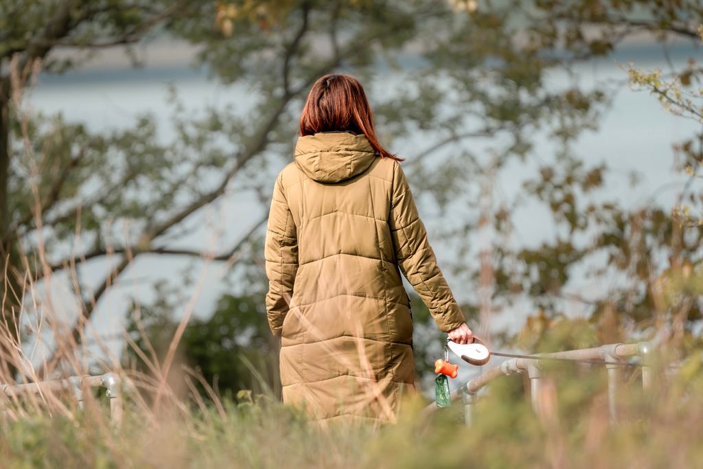 Woman in a green coat strolls through a scenic autumn park with trees and a lake.